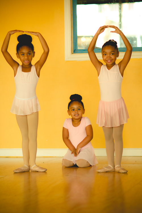 Three young ballet students in white leotards and skirts. Two girls stand in first position with their arms above their head.